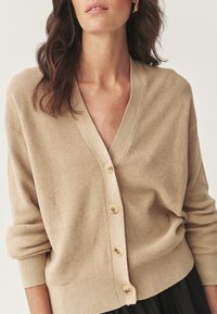 Femme portant un cardigan en tricot beige à boutons avec des manches longues retroussées, associé à une jupe noire, montrée des épaules aux hanches.
