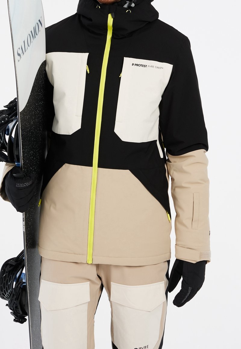 Personne portant une veste et un pantalon de ski beige, noir et blanc tenant un snowboard Salomon noir et blanc avec des gants noirs.