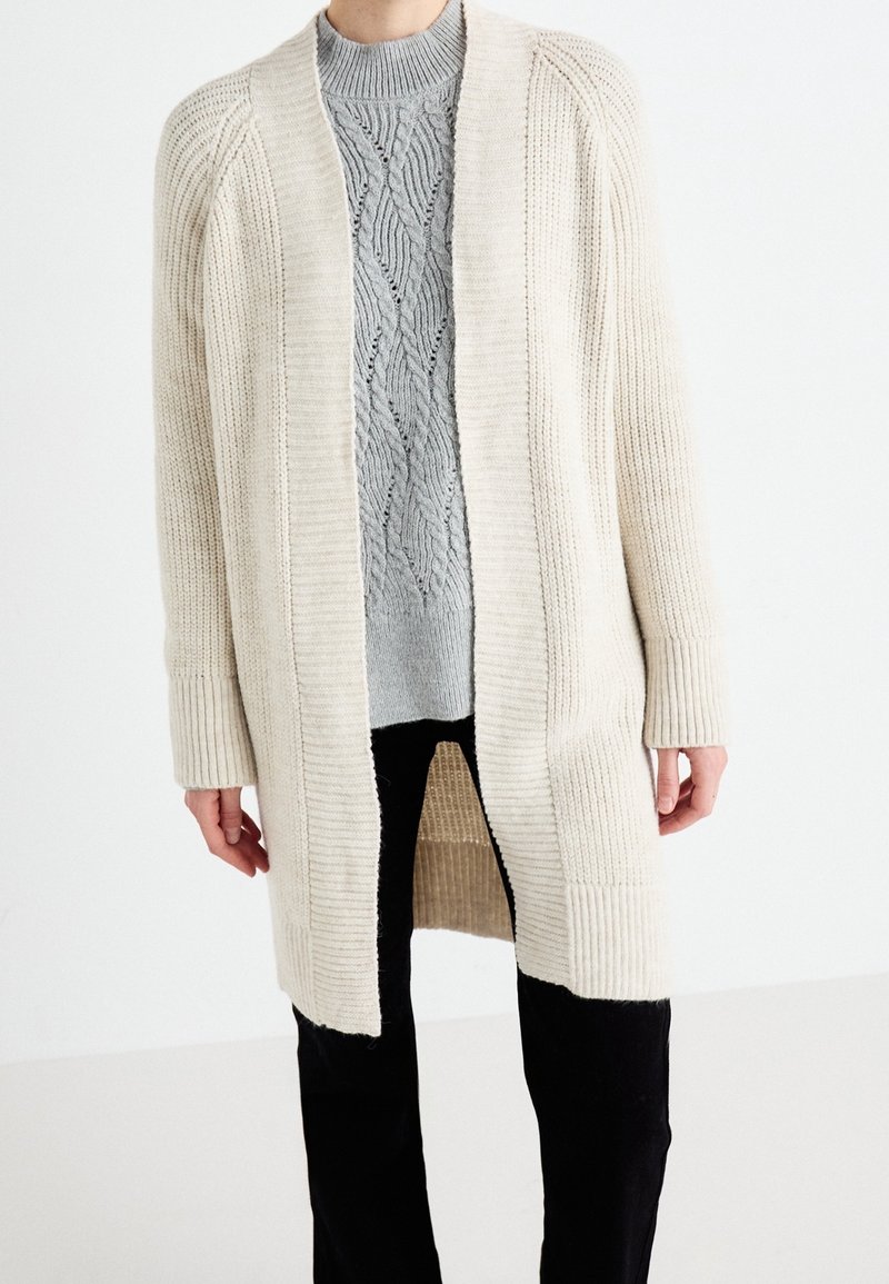 Personne portant un long cardigan côtelé crème sur un pull en maille gris à motifs et un pantalon noir, debout devant un fond blanc.