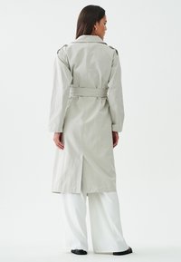 Heled hall trench coat, millel vööga, pikad varrukad ja seljatagune vent. Sobitatud laiade valgete pükste ja mustade kingadega.
