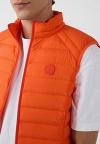 Oranje gewatteerd vest met hoge kraag, ritssluiting, textuur rijen en een ronde logo aan de voorkant. Draagt over een wit t-shirt.