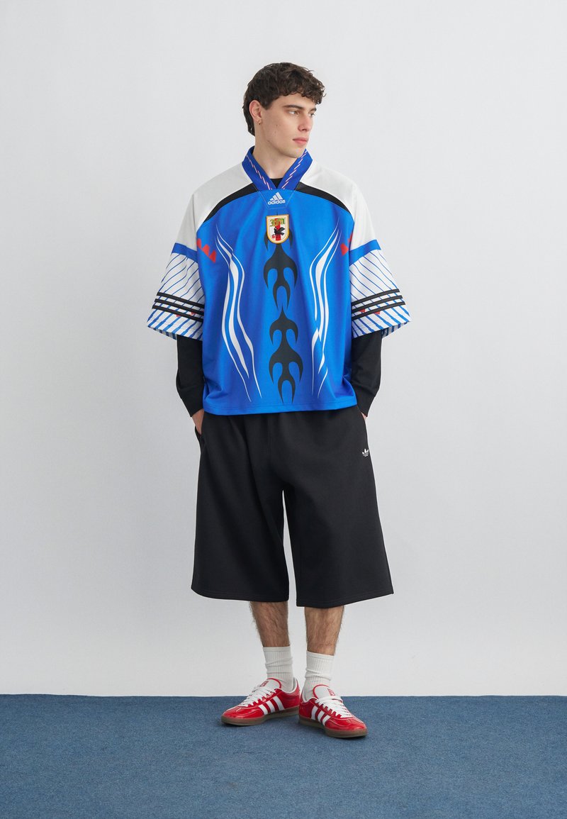 Jeune homme portant un maillot de sport oversize à motifs bleu, blanc et noir, un short large noir, des chaussettes blanches et des baskets rouges Adidas.