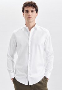 Weißes Langarmhemd mit Button-Down-Kragen, taillierter Passform und glatter Textur; kombiniert mit khakifarbenen Hosen.