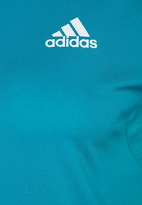 Turkoosi urheilupaita, joka on valmistettu sileästä kankaasta ja jossa on valkoinen Adidas-logo vasemmassa yläkulmassa. Minimalistinen muotoilu, lyhyet hihat.