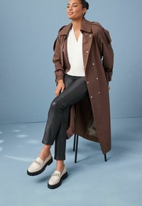 Ruskea trench-viittapaitaan päällä, valkoinen villapaita, mustat nahkahousut ja beiget loafers-kengät, joissa on ketjustyyliset koristeet, sinistä taustaa vasten.