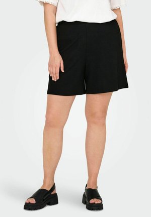 Mujer con pantalones cortos negros de talle alto y sandalias negras robustas, con las manos relajadas a los lados, sobre un fondo liso y claro.