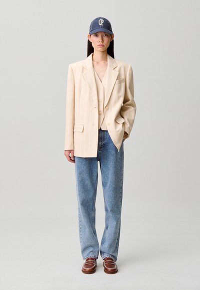 Blazer beige a righe sottili sopra un gilet crema, abbinato a jeans a gamba larga azzurri e mocassini marroni. Un berretto blu scuro completa il look.