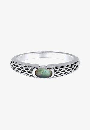 Anillo de plata con diseño trenzado y acento de turquesa, llevado en una mano que descansa en un bolsillo, contra un fondo de vaqueros negros y una camisa blanca.