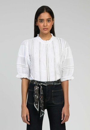 Blouse blanche à manches bouffantes avec des rayures verticales, dentelle, et col festonné, associée à un jean foncé et une ceinture à motif en écharpe.