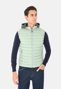 Uomo con gilet imbottito verde chiaro e cappuccio grigio sopra una maglietta a maniche lunghe blu navy e jeans blu scuro, in posa con una mano in tasca.