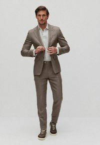 Beżowy garnitur o dopasowanym kroju, zestawiony z jasnoniebieską koszulą w paski i ciemnobrązowymi tenisówkami. Spodnie szyte na miarę oraz eleganckie detale stylizacji.