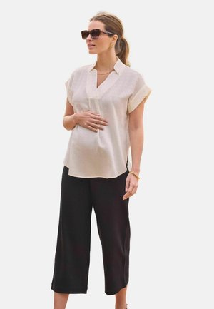 Terhes nő, bézs texturált rövid ujjú blúzban, fekete, széles szárú, rövidített nadrágban, arany ékszerekkel és teknőspáncél mintás napszemüveggel.