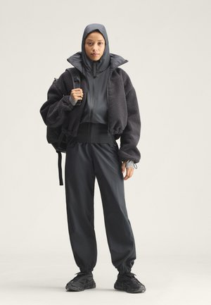 Young person wearing black hooded jacket, loose black pants, black sneakers, and carrying a black backpack, standing against a plain background.