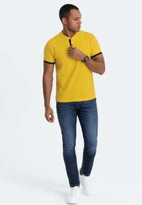Kortærmet gul polo-skjorte med sorte detaljer og en stolpe. Båret med mørkeblå jeans og hvide sneakers. Simpelt, tætsiddende design.