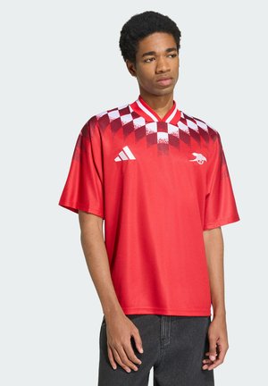 Młody mężczyzna noszący czerwoną i białą sportową koszulkę w kratę z logo Adidas i Arsenal, stoi z neutralnym wyrazem twarzy.