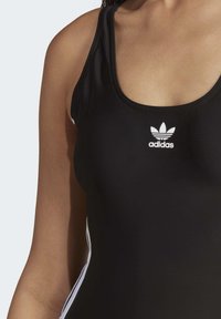 Črn atletski kopalke s krožnim izrezom, ki ima bele stranske črte in majhen bel Adidasov logotip na prsih. Gladek tekstilni material.