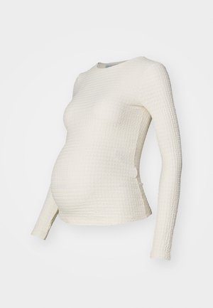 Langermet gravidebluse i strukturert kremfarget stoff, med en tettsittende form og rund hals, designet for å romme en voksende mage.
