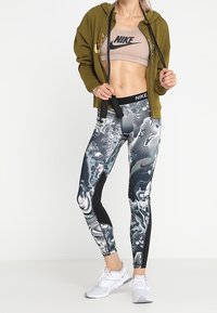 Kvinna som bär en olivgrön zip-up hoodie, beige Nike sport-bh, svarta och vita mönstrade leggings, vita sneakers och håller i svarta hopprep-handtag.