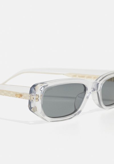 Occhiali da sole con montatura trasparente e lenti scure, con bracci texture dorati e resistenti cerniere. Forma moderna con design angolare prominente.