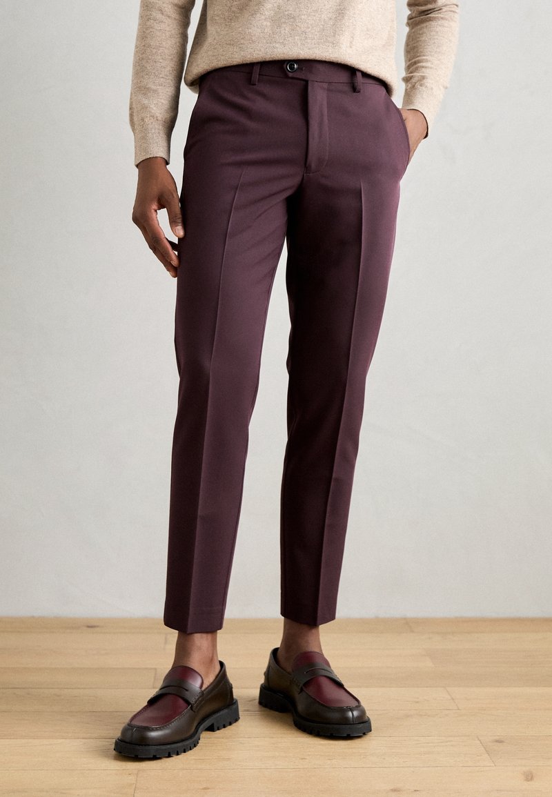 Personne portant un pantalon slim fit bordeaux foncé avec des plis devant et des mocassins en cuir marron foncé, debout sur un sol en bois.