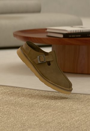 Slip-on sko i olivengrønt semsket skinn med metallspenne og tykk lys brun såle, plassert over et beige teppe nær et rundt trebord.