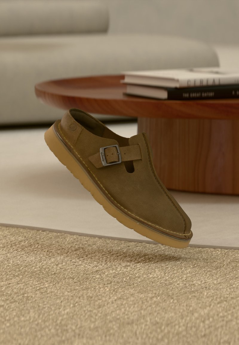 Πράσινο ελιάς σουέτ παπούτσι τύπου slip-on με μεταλλική πόρπη και χονδρό καφέ τακούνι, τοποθετημένο πάνω από μπεζ χαλί κοντά σε στρογγυλό ξύλινο τραπέζι.