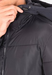 Giacca a vento nera con zip frontale, cappuccio regolabile e una texture liscia e lucida. Presenta cuciture visibili e accessori metallici.