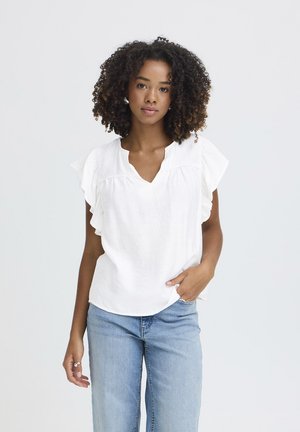 Ung kvinde med krøllet hår iført en hvid kortærmet bluse og lyseblå jeans, stående mod en ensfarvet hvid baggrund.