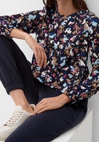 Blusa casual di un blu scuro con motivo astratto multicolore, caratterizzata da maniche lunghe a sbuffo e scollatura rotonda. Abbinata a pantaloni blu.