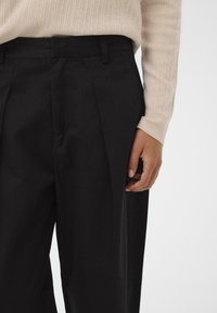 Schwarze maßgeschneiderte Hosen mit Falten, aus einem glatten Stoff gefertigt. Ein hellbeiges geripptes Sweatshirt ist getragen und teilweise links sichtbar.