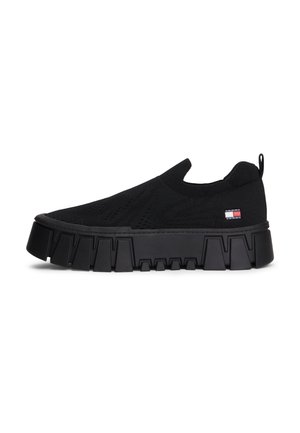 Musta slip-on-tennari paksulla, massiivisella pohjalla ja pienellä punaisen, valkoisen ja sinisen lipun logolla sivussa, neulottu päällinen.