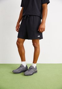 Homme portant un short noir New Balance, un t-shirt noir, des chaussettes blanches et des baskets grises New Balance, debout sur un sol vert.