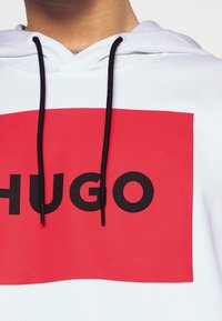 Weißer Hoodie mit einem großen roten rechteckigen Grafik und dem schwarzen Text "HUGO." Schwarze Kordeln sind am Kragen sichtbar.