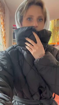 Casaco puffer preto acolchoado com colarinho alto, apresentando um cinto e tecido texturizado. Usado com um hoodie branco suave por baixo.
