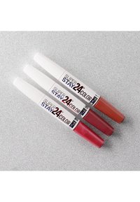 Tre tubetti di rossetto con cappucci bianchi, disponibili in tonalità di rosso e nude. Le etichette riportano la scritta "SUPER STAY 24 COLOR" su uno sfondo argentato.
