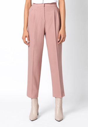 Femme portant un pantalon rose poudré taille haute, coupe droite, associé à des bottines beige clair à bout pointu, debout devant un fond neutre.