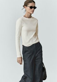 Top crema a maniche lunghe con scollo rotondo, abbinato a pantaloni denim scuri a gamba larga. Il modello indossa occhiali da sole rettangolari scuri e tiene una borsa nera.