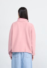 Pull en polaire rose avec un col monté, des épaules tombantes et une poche carrée au dos. Tissu doux et texturé ; coupe décontractée.