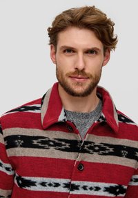 Red and beige plaid shirt jacket with black and gray patterns, featuring a collar, front buttons, and a textured gray sweater underneath.