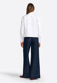 Blouse blanche à col avec un décolleté volanté ; associée à un pantalon en denim bleu foncé à jambes larges avec poches arrière et un ourlet soigné.