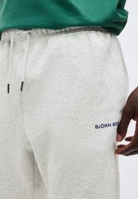 Κοντινό πλάνο ατόμου που φοράει ανοιχτό γκρι φόρμα με κορδόνι και λογότυπο "BJÖRN BORG" στο μηρό, σε συνδυασμό με πράσινο τοπ.