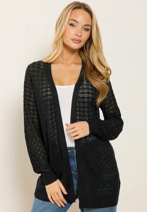 Lyshåret kvinde iført en sort tekstureret cardigan med åbent front over en hvid top og blå jeans mod en ensartet lys baggrund.