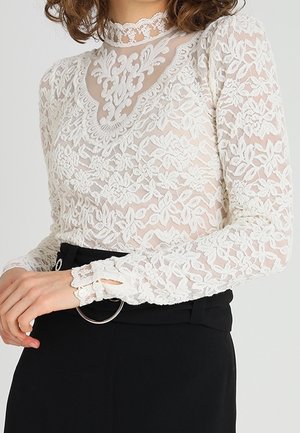 Femme portant un haut blanc en dentelle florale à col montant et manches longues, associé à un pantalon noir taille haute et une ceinture argentée à boucle ronde.