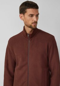 Bordeauxroter Reißverschluss-Pullover aus strukturiertem Stoff mit einem geometrischen Muster. Hoher Kragen, Rippdetails und ein kontrastierender Reißverschluss.
