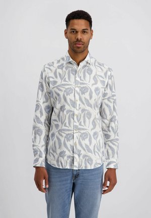 Mann mit weißem Hemd mit grauem Blumenmuster und blauer Jeans, vor schlichtem hellem Hintergrund stehend.