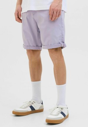 Shorts violets clairs avec un ourlet roulé, en tissu lisse. Portés avec des baskets blanches avec des rayures marines et des chaussettes blanches. 