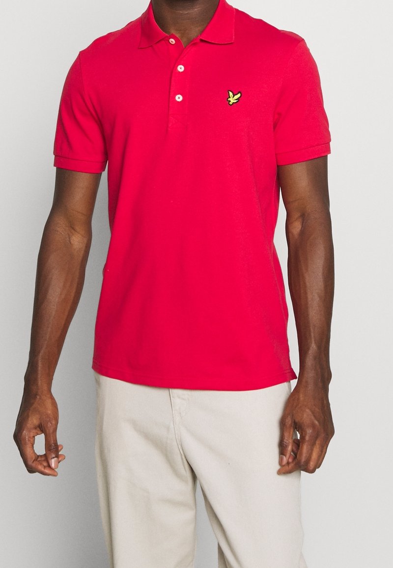 Homme portant un polo rouge vif à manches courtes avec un logo oiseau jaune et un pantalon beige clair sur un fond uni.