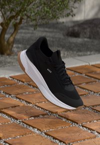 Sneaker BOSS nera con suola bianca, lacci e suola in gomma, sospesa sopra piastrelle di legno all'aperto con sfondo sfocato di alberi e pietre.