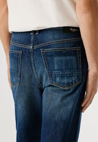 Denimové džíny v tmavě modré barvě s hladkou texturou, s zadními kapsami a viditelným švem a černou značkovou visačkou na pase.