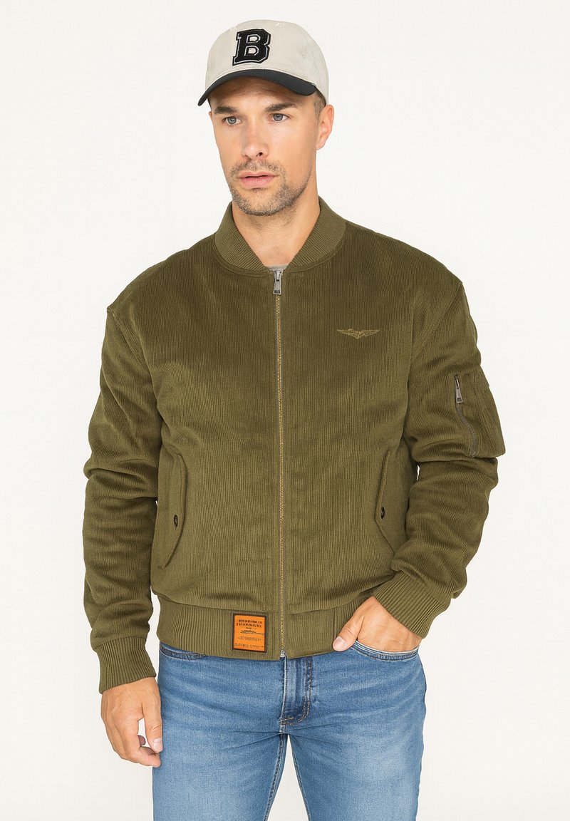 Veste bomber en velours côtelé vert olive avec fermeture éclair à l'avant, poignets côtes, deux poches latérales et un logo sur la poitrine. Portée par-dessus un jean bleu.